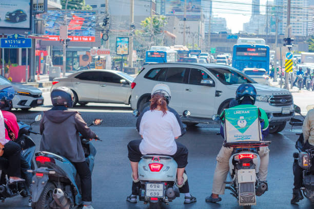 Waiting thai people on motorcycles at junction of Rama 9 Road in Bangkok Huai Khwang / Din Daeng. People arec standing in shadow of overpass road over junction from east to west. People are standing ahead of stoplight among lines into both directions. Cars are crossing the junction from left to right. In background is queued traffic jam on Thanon Asok towards Sukhumvit Road