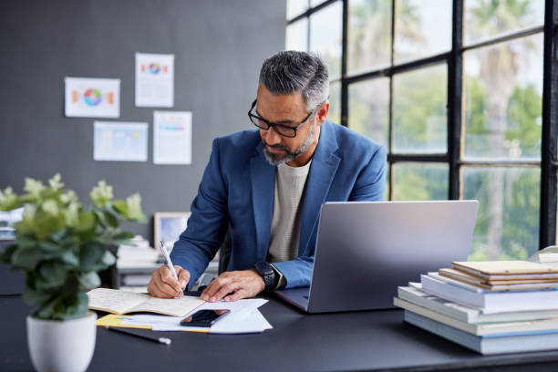 Middle eastern business man in blue blazer working at desk with laptop while taking notes. Mid adult professional man writing in notebook in office. Thoughtful smart casual businessman reviewing documents in modern workspace with copy space.