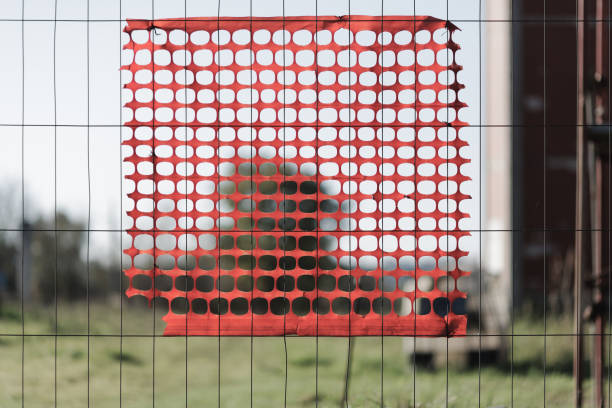 A close-up view of a construction site feature, showcasing a section of red safety mesh attached to a metal fence. The vibrant red contrasts with the blurred green landscape in the background, creating an abstract and unconventional perspective of a construction site. The composition highlights textures, shapes, and the interplay of industrial materials with natural surroundings