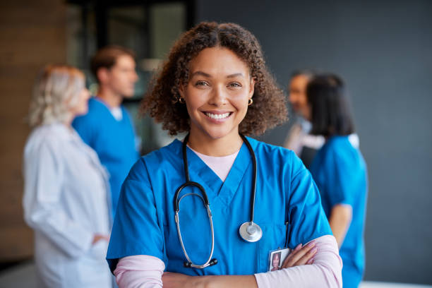 Portrait of smiling african woman nurse Confident happy woman nurse smiling with crossed arms with healthcare team in background. Multiethnic medical professional in scrub ready to assist patients. Portrait of reliable confident nurse standing proudly, surrounded by colleagues in a private clinic. nurse stock pictures, royalty-free photos & images