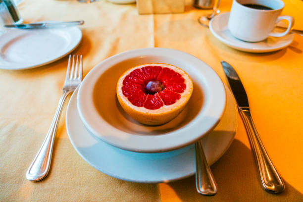 A simple breakfast plate featuring a halved grapefruit topped with a single blueberry. The table is set with silverware, coffee, and plates on a soft yellow tablecloth.