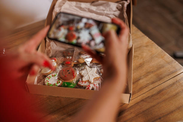 A person captures a photo of festive Christmas cookies in a cardboard box, highlighting holiday joy and creativity. Vibrant decorations and rustic packaging convey warmth and seasonal celebration.