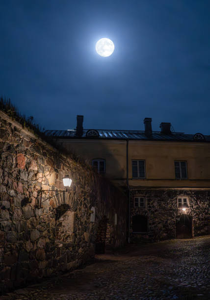 A moonlit view into an area of the large sprawling Suomenlinna Castle, a UNESCO world heritage site, on the eponymous island in front of Helsinki, Finland. In the photo a stone wall can be seen, topped by a residential building, the scene illuminated by old-fashioned lanterns and full moon