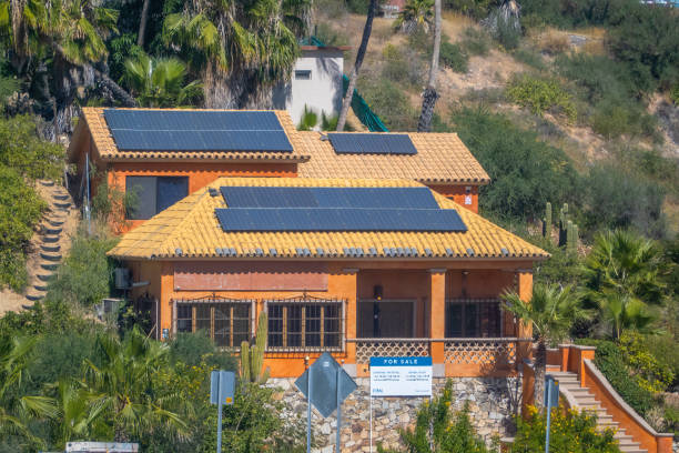 Los Cabos, B.C.S., Mexico. Nov 22, 2024. A multi-level orange house with solar panels, a For Sale sign, and a palm tree-lined hillside backdrop.