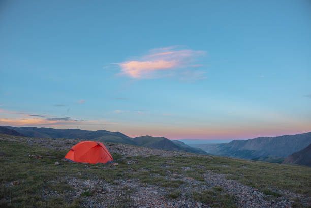 istock Orange tent on grassy stony pass with view to valley and mountain range silhouette with snowy peaks of vivid pink color far away at sunset. Red tent in high mountains under cloud illuminated by sunset 2187643101