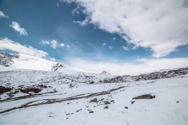 istock Cold snow-white landscape with snow-covered pointy peak under clouds in blue sky. Snowy rocky mountainside and mountain peaked top. Last year's grass and stones in snow and sharp pointed pinnacle. 2187643127