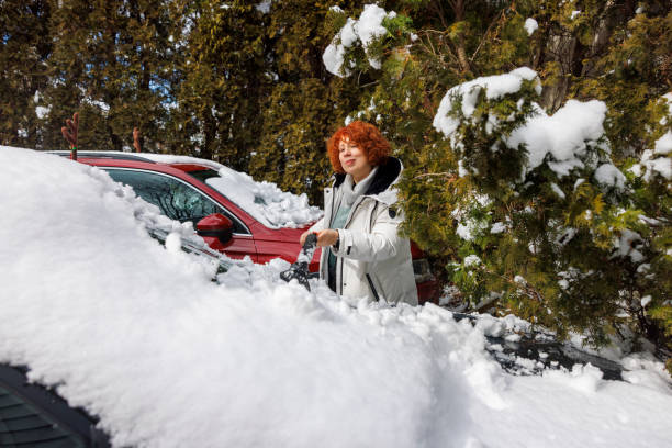 Winter car care: red-haired woman clears snow from her car in the yard using a scraper