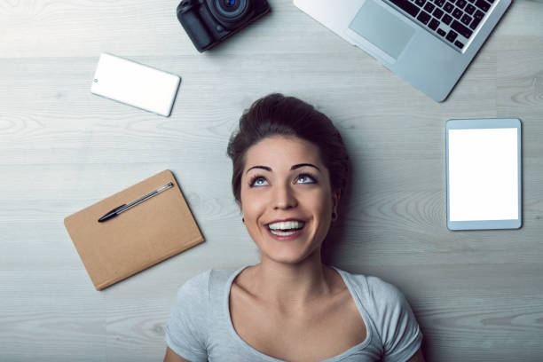 istock Young woman lying on the floor smiling and looking up, surrounded by technology 2187654582