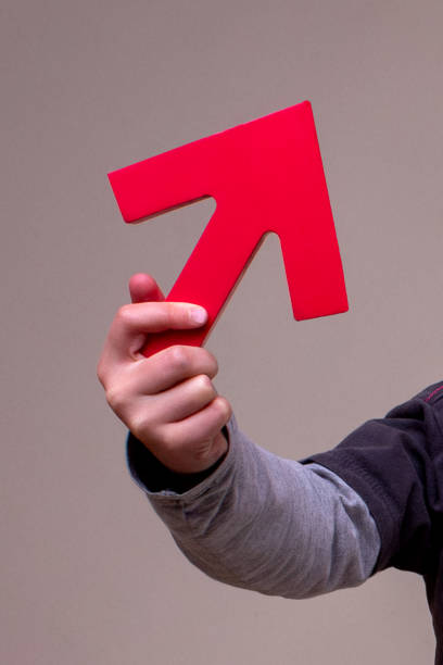 Hand holds big red arrow pointing upwards.  closeup of hand holding red arrow pointing upward in gray background