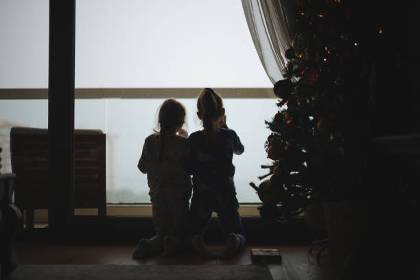 Two cute children sitting by window on Christmas eve. Two school toddler girls, siblings looking outdoor and dreaming. Family happiness on traditional holiday