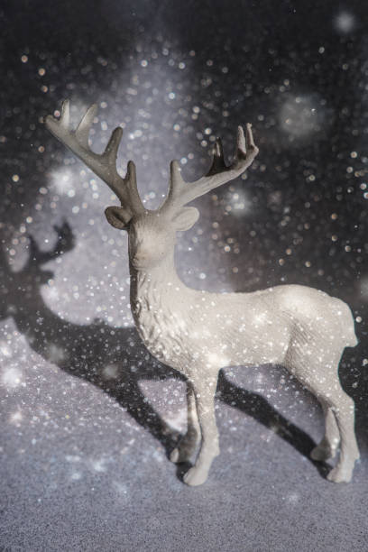 A close-up shot of a golden glittering reindeer ornament placed in front of a decorated Christmas tree. The festive holiday setting features beautiful pastel-colored baubles, pink bows, and a soft snowy background, creating a magical atmosphere perfect for the Christmas season.