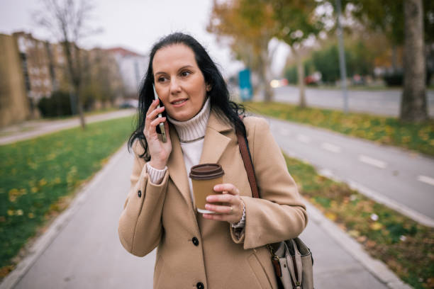 istock A middle-aged woman in a stylish coat, holding a takeaway coffee cup in one hand while talking on her phone with the othe 2187720950