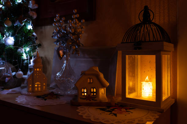 A warm and festive Christmas setup featuring a glowing lantern, ceramic houses with lighted windows, and a small Christmas tree decorated with ornaments. The soft, ambient lighting creates a cozy and inviting holiday atmosphere.