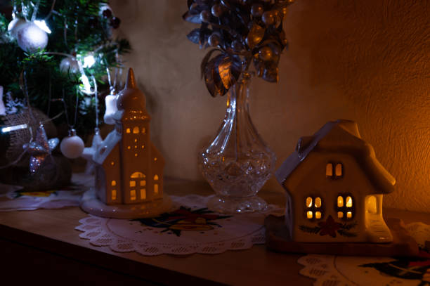 A charming Christmas display featuring illuminated ceramic houses and a glass vase with decorative silver berries. Soft, glowing light creates a warm and festive ambiance, complemented by a small Christmas tree adorned with ornaments in the background.