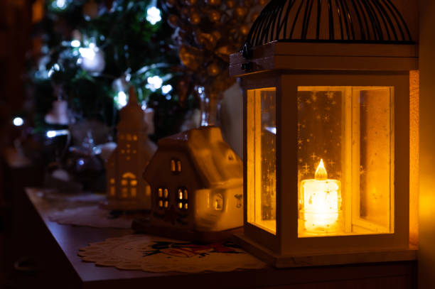 A warm and inviting Christmas scene featuring a glowing lantern, illuminated ceramic houses, and a beautifully decorated Christmas tree in the background. The soft candlelight and twinkling tree lights create a magical and cozy holiday atmosphere.