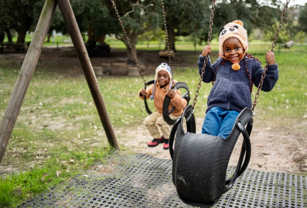 Smiling little girl playing with her cute baby brother on a swing outside in a playground during winter