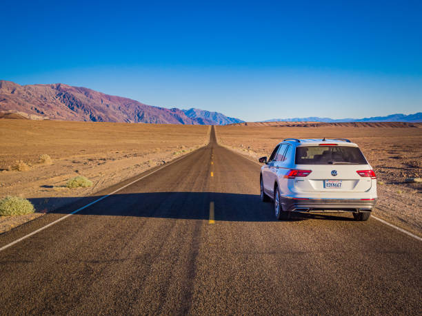 White suv is driving down a long, straight road in the desert, heading towards a range of mountains in the distance under a clear blue sky