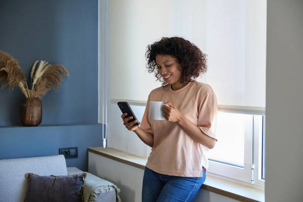 istock Latin woman using smartphone and drinking coffee by the window at home 2187751151