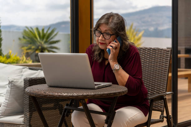 istock Elderly woman talks on her cell phone while looking at her laptop on a terrace 2187753005