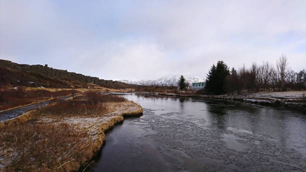 Iceland - 3.24.2018: A frozen river through dried meadows next to a forest and cliffs with snow mountains at the horizon under a cloudy sky in Thingvellir National Park before the pandemic