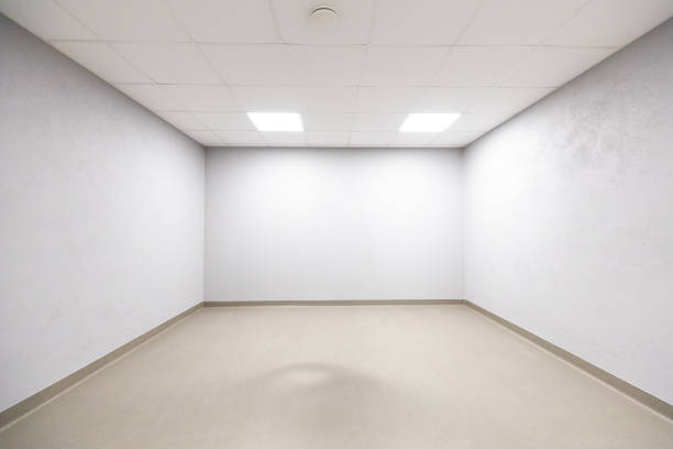 An empty room with plain white walls and a beige floor. The ceiling features a grid of recessed lighting panels, creating a bright, open space.