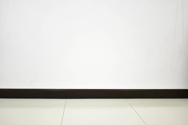 A clean white wall with a dark wooden baseboard and light tiled flooring, ideal for backgrounds or minimalist designs.
