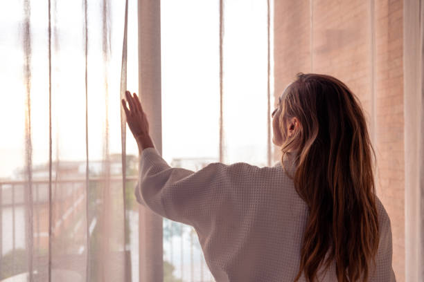 istock Woman opening curtains in a sunlit room with a view 2187766861