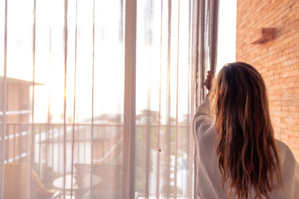 istock Woman opening curtains in a sunlit room with a view 2187766862
