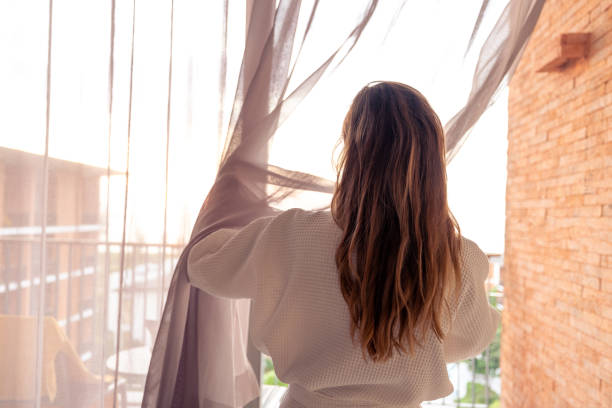 istock Woman opening curtains in a sunlit room with a view 2187766867