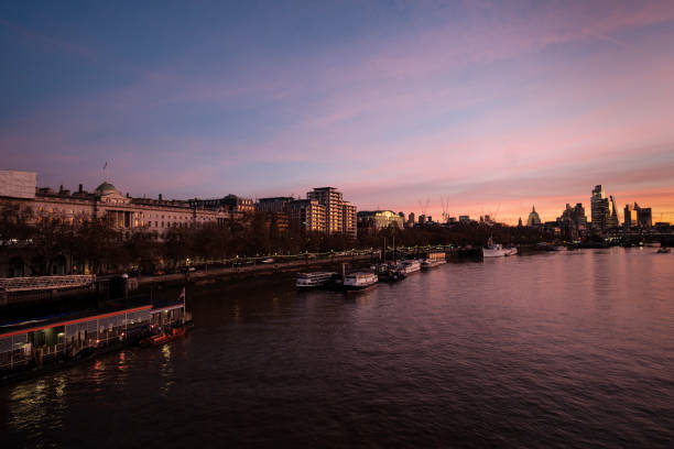 Colorful sunrise illuminates buildings along the thames river in london, england, with moored boats adding to the picturesque urban scene