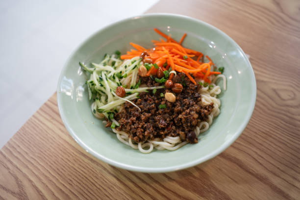 Chinese noodles topped with minced meat in black sauce, garnished with cucumber slices, carrot slices, and peanuts.