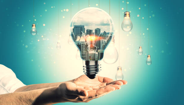 istock Lightbulb with urban skyline within, supported by hands on teal background. 2187792271