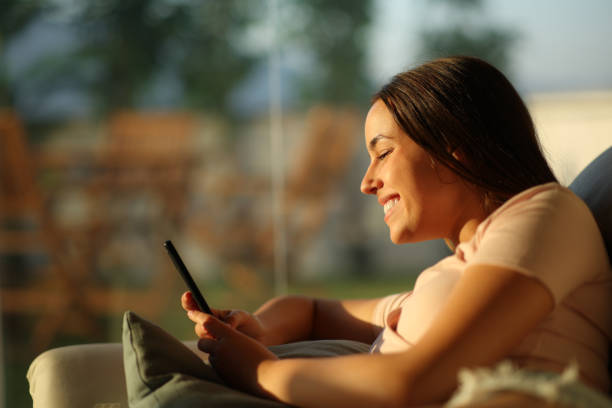 Happy woman using phone at sunset at home stock photo