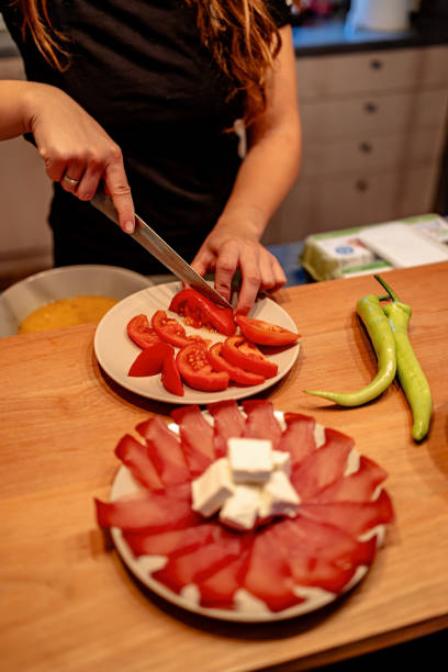 In a bright kitchen, a young woman cuts red tomatoes with a knife, arranging them on a plate while vibrant green peppers rest nearby, preparing for cooking.