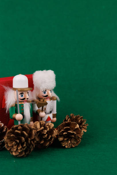 A whimsical wooden nutcracker and a teddy bear are placed next to a red gift, surrounded by silver baubles and decorative pine cones on green