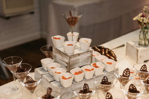 Luxurious catering setup featuring rows of chocolate mousse served in martini glasses decorated with chocolate garnishes, alongside white ceramic cups with orange-colored desserts. A beautiful wildflower arrangement with purple asters and seasonal flowers adds a rustic touch to the elegant dessert table. The presentation is set on a cream-colored tablecloth in an indoor venue, perfect for wedding receptions, corporate events, or upscale celebrations.