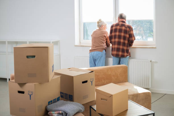Senior couple moving house Heap of cardboard boxes with senior couple looking through window in the background. Senior couple moving house. seniors-moving-house stock pictures, royalty-free photos & images