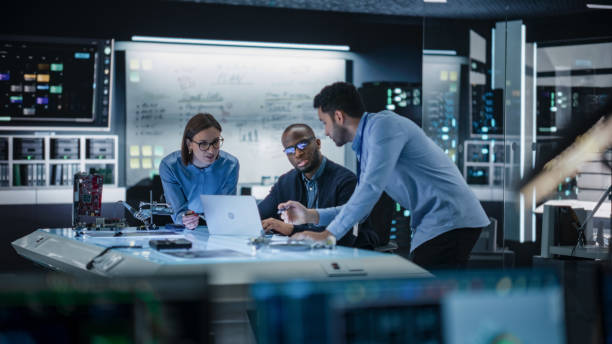 istock Diverse Team of Engineers Collaborating on Manufacturing Project in a Research Facility. Colleagues Analysing Data Using Laptop Computer and Brainstorming for a Technologically Advanced Invention 2187841576