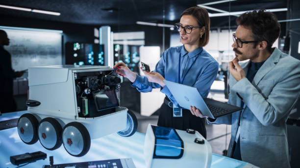 istock Portrait of Two Colleagues Working in a Research Facility, Using Laptop Computer to Develop a Futuristic Robot for Autonomous Deliveries. Engineers Working on Improving Detectors on their Prototype 2187842704