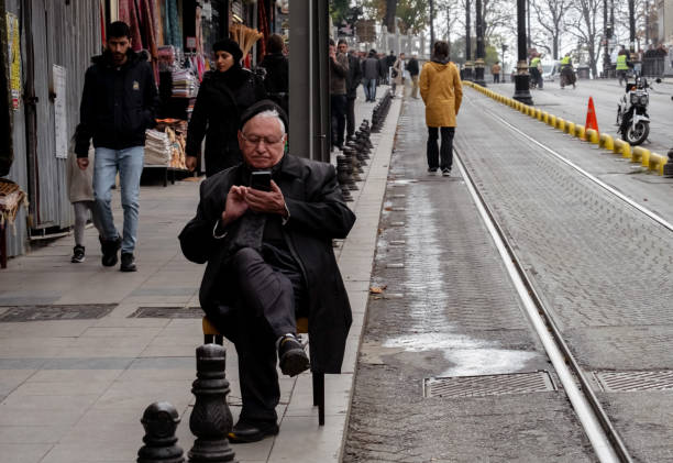An elderly man is sitting at a tram stop and using a mobile phone stock photo