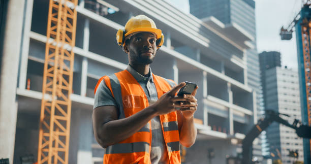 istock Portrait of Handsome African Builder, Worker, Craftsman Wearing a Hard Hat and a Vest while Standing at a Commercial Building Construction Site and Using a Smartphone to Browse Through Social Media 2187851391