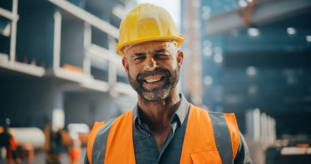 istock Portrait of Happy Successful Construction Worker Standing in a Construction Yard, Posing for Camera, Smiling. Professional Civil Industry Engineer Wearing Safety Uniform and Protective Hard Hat 2187851835