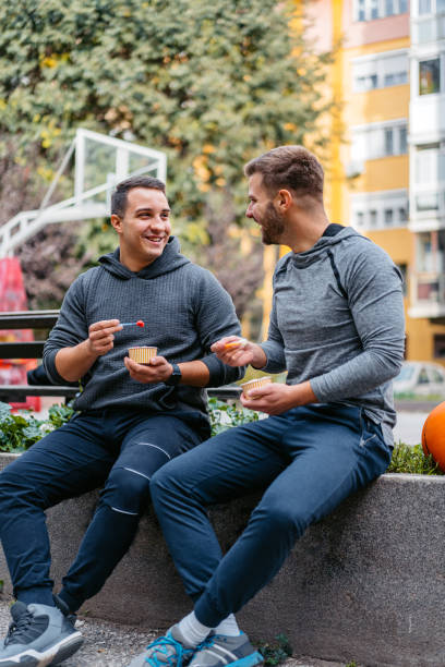Two senior multi-ethnic men sitting outdoors together on a park bench. The Caucasian man, in his 80s, has h is arms around his friend's shoulders and they are smiling.
