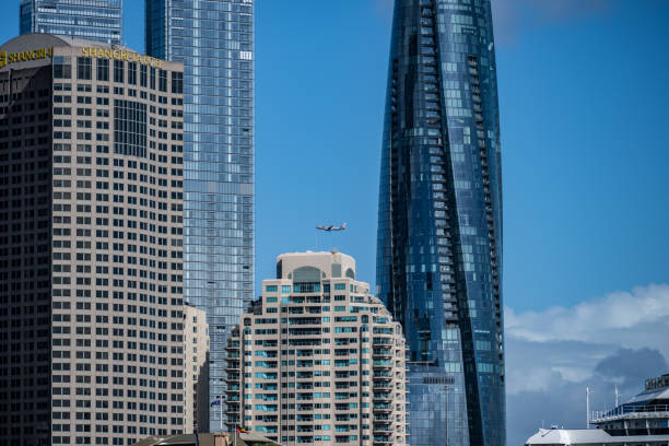 A plane flies above Sydney’s skyline, featuring modern skyscrapers like the Crown Tower and Shangri-La Hotel against a clear sky.