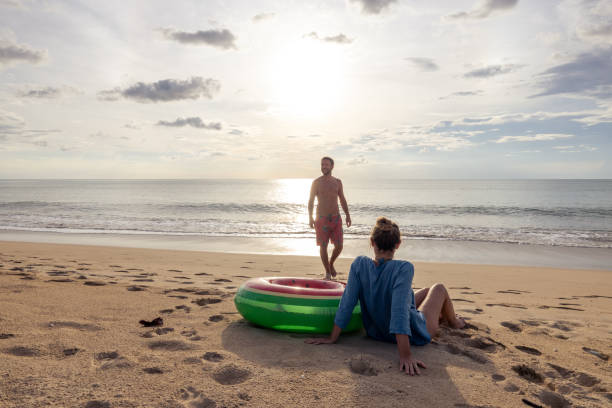 istock Man standing on sandy beach while woman relaxes nearby with inflatable ring at sunset 2187857438