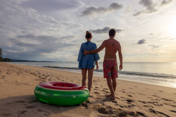 istock Couple Standing on Beach at Sunset Embracing Next to Inflatable Pool Ring 2187857673