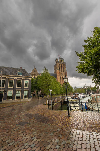 istock Historic European town with a clock tower under a dramatic cloudy sky. Cobblestone streets lead to a marina filled with boats. Trees and old architecture frame the peaceful scene. Dordrecht landscape 2187863402