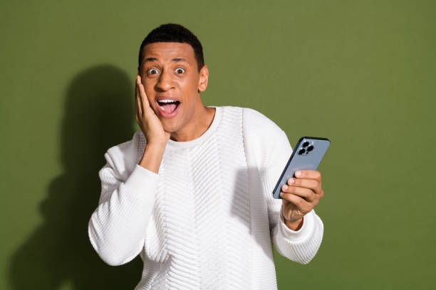 istock Young man in white sweater expressing excitement while looking at smartphone against khaki background 2187868964
