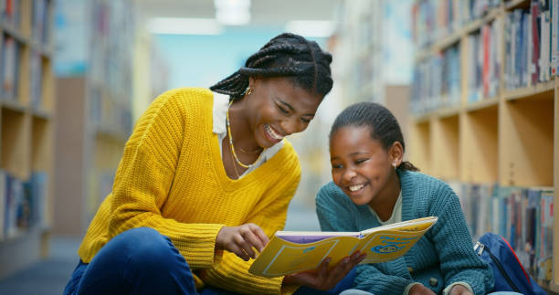 istock Library, child and teacher with book for laughing, learning and knowledge for storytelling and language. Educator, black woman and girl on floor for fun, fantasy stories and development or relaxing 2187876923