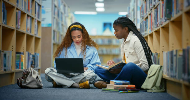 istock Education, students and women in library, laptop and teamwork for assignment, research and internet. People, university and friends on floor, computer and conversation with network, tech and learning 2187876984